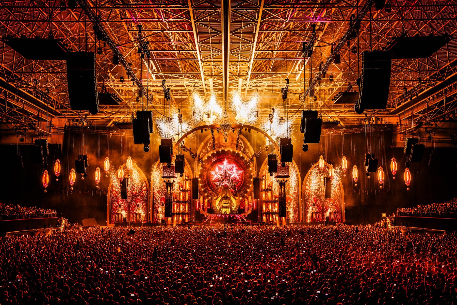 Pyro fountains and fireworks behind an ornate arena stage in front of a packed crowd