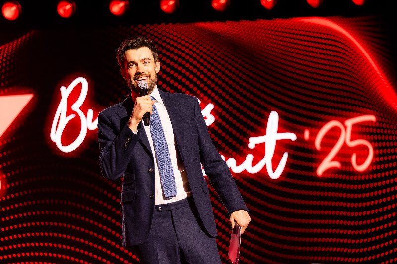 F1 Presenter in a navy suit speaking into a microphone in front of a red LED screen with neon lettering