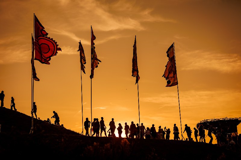 Silhouettes of festival-goers walking up a hill past tall red flags at sunset