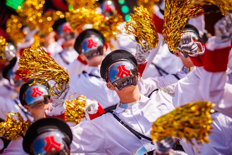 Performers in white uniforms, black berets and mirrored face masks with a red logo waving gold pom-poms