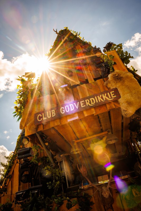 Wooden DJ cabin with a Club Godverdikkie sign and sheepskins, shot upward into the sun with a lens flare