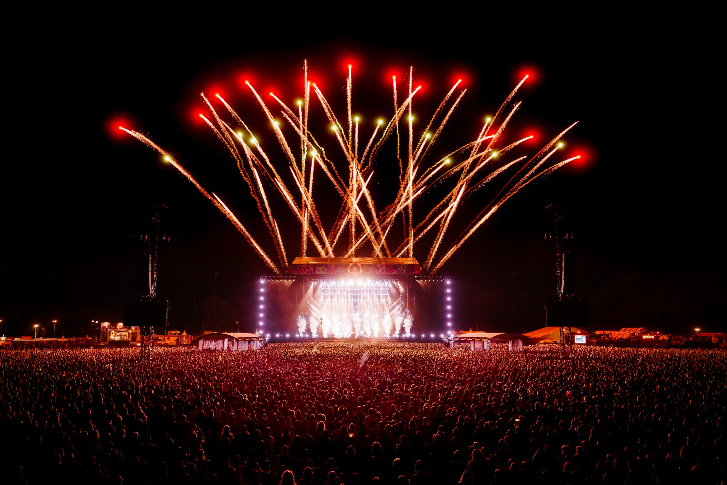 Red and gold firework fountains above a main stage in front of an enormous night-time crowd
