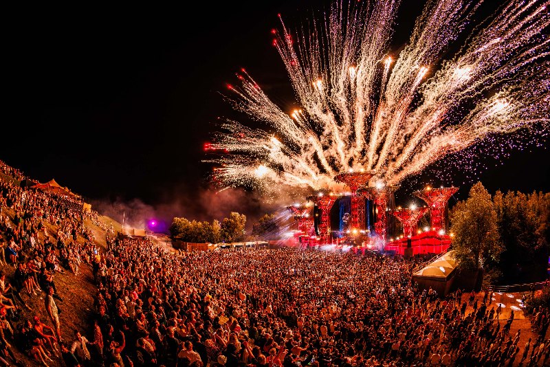 Firework burst above a red-lit stage seen from a hillside crowd at night