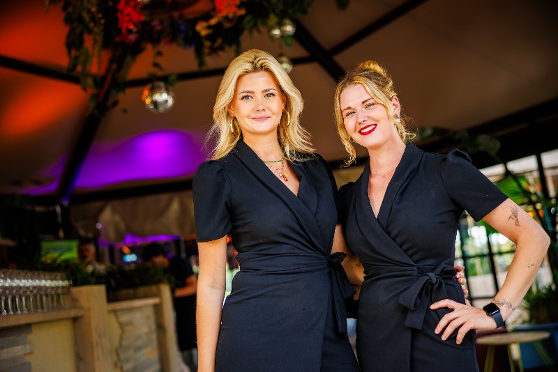 Two hostesses in black wrap dresses smiling in a decorated hospitality tent
