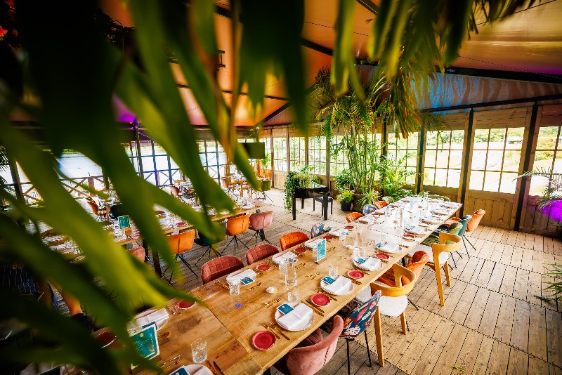 Long wooden dining tables set with colourful plates in a lakeside pavilion surrounded by palms
