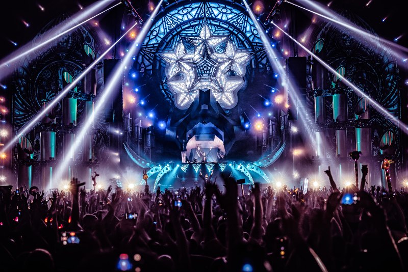 DJs with raised arms on a stage with a giant white star and stained-glass design, light beams cutting through haze over a crowd