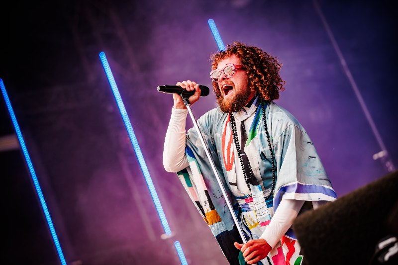 Singer with curly hair and red glasses performing into a microphone in purple stage light