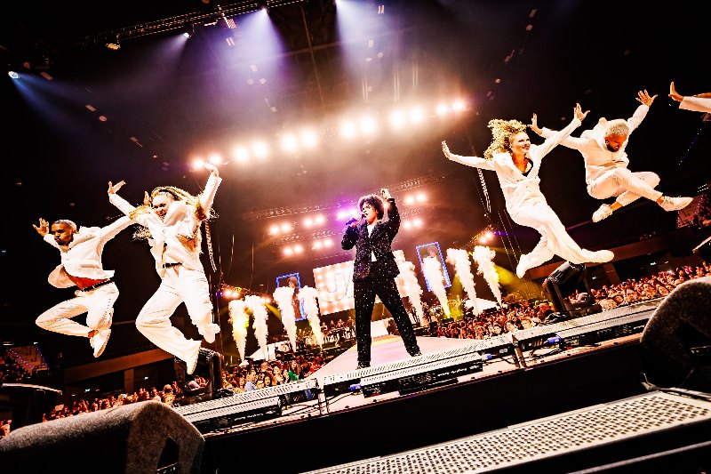 Singer on stage with dancers in white suits leaping mid-air in front of white smoke jets