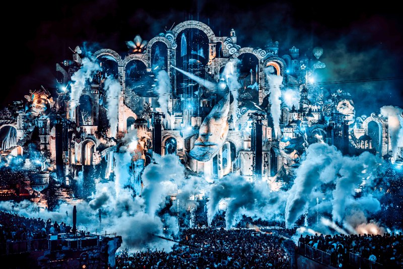 Mainstage with giant stone faces and arches wrapped in blue smoke and light, with a crowd below