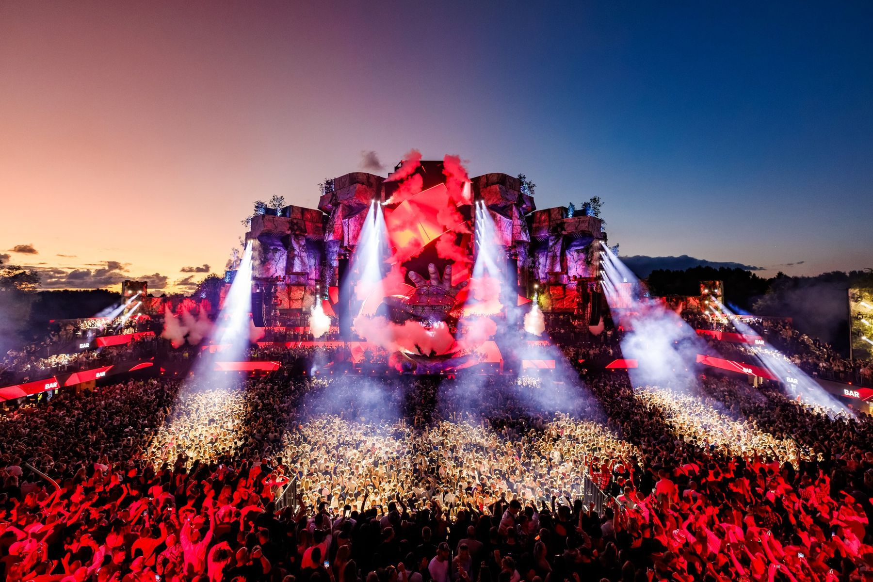 Mysteryland main stage at sunset, a giant stone hand holding the DJ booth, red smoke and light beams over the crowd